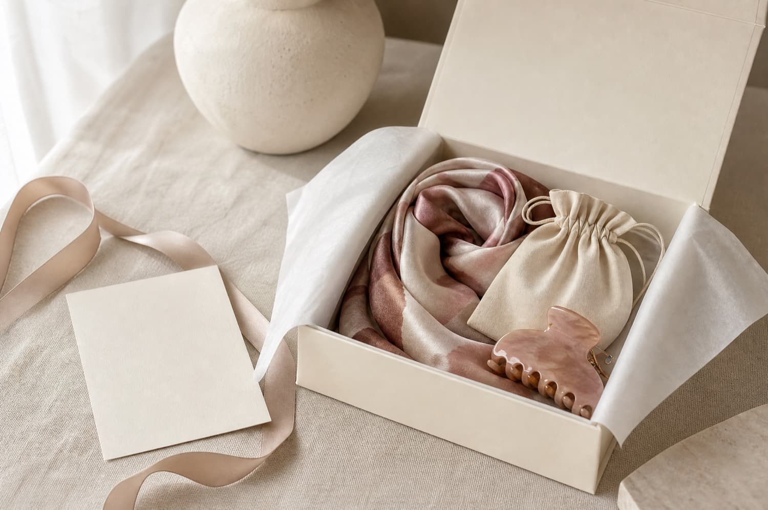 An open cream gift box with tissue folded back revealing a rolled scarf and a small pouch
