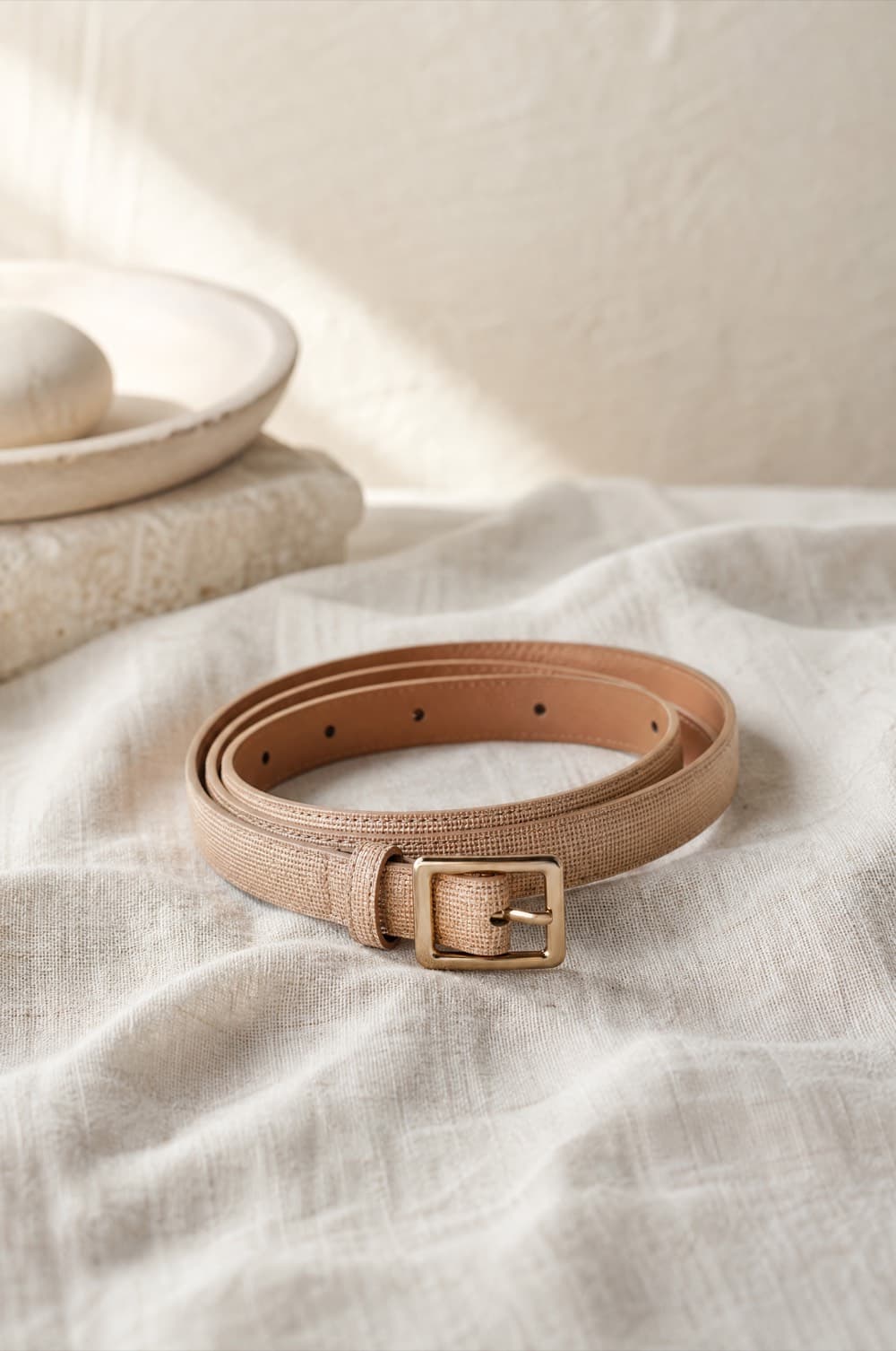 Narrow tan belt with a small square gold-tone buckle coiled on linen